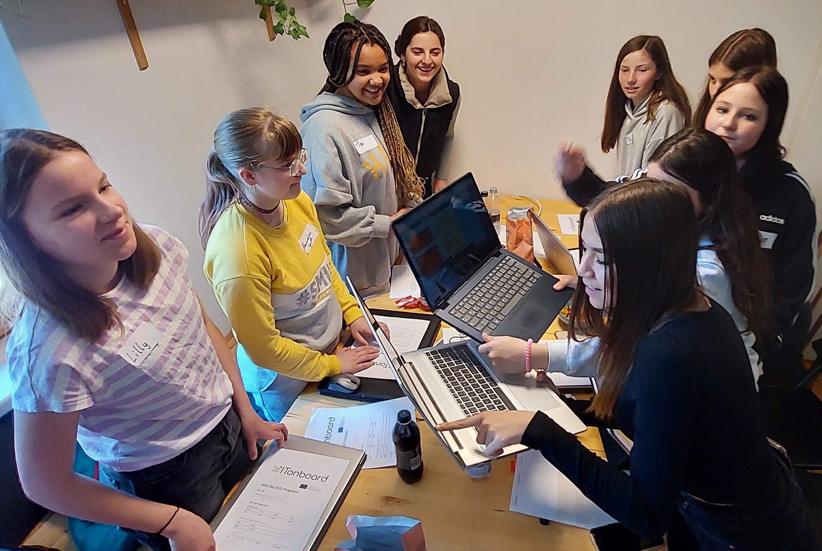 9 girls are standing around a conference table holding computers in their hand and reading from them while talking.