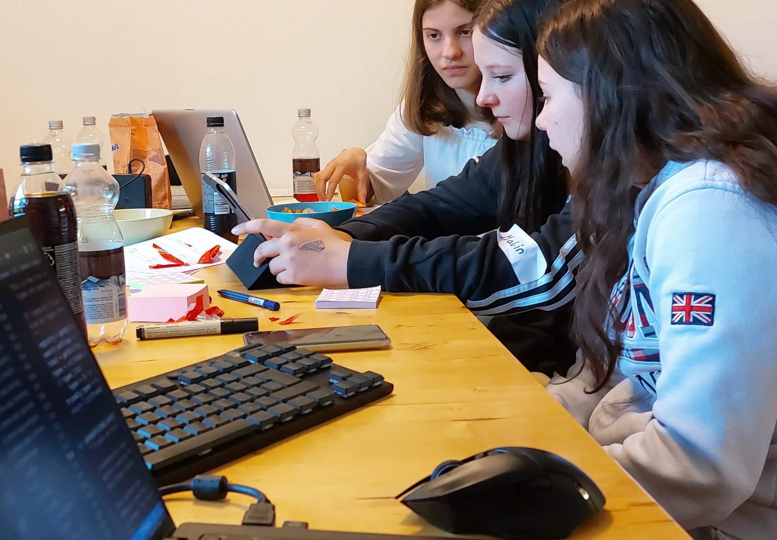 3  girls on a table with sweets and soft drink bottles sitting in front of their computers watching their screens while discussing and writing or drawing on Post Its.